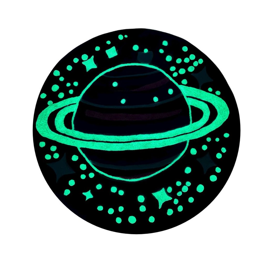 Handmade tufted rug Cosmos glowing in the dark, showing illuminated Saturn design with blue and purple tones.